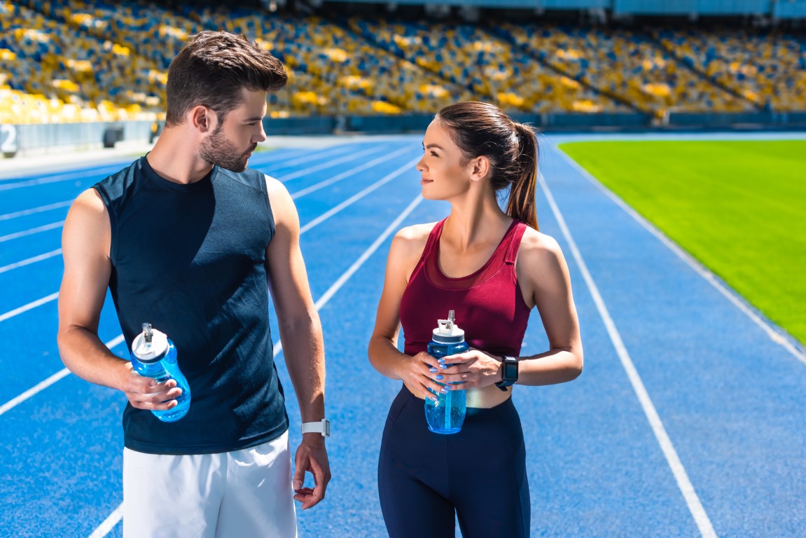 young sportive couple with bottles of water chatting on running track at sports stadium